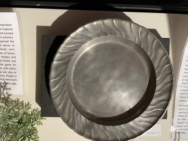 A plate made of pewter, with a central dish and scalloped edges, on display in a museum case.