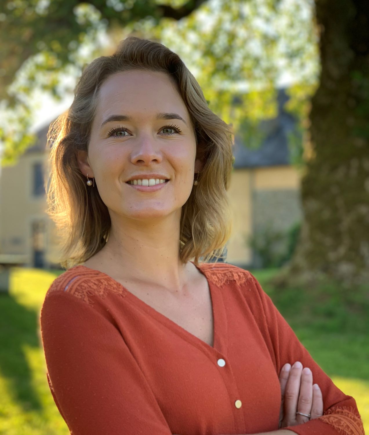 Portrait of Iris de Rode. She has blonde hair and is wearing a salmon colored shirt. She is standing outside on a sunny day.