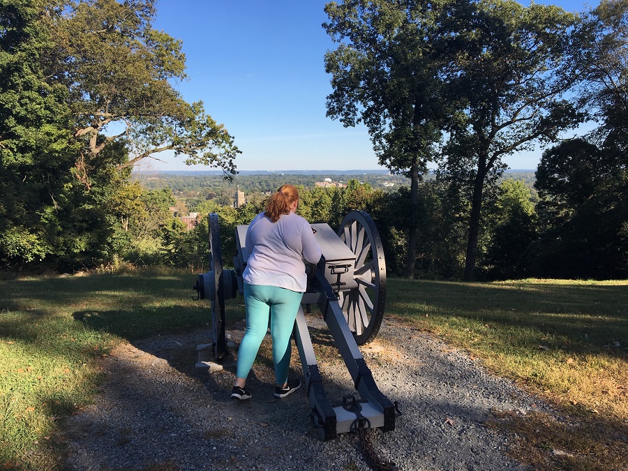 A woman standing near a cannon which is a an overlook called Fort Nonsense.  The town of Morristown is on the horizon.