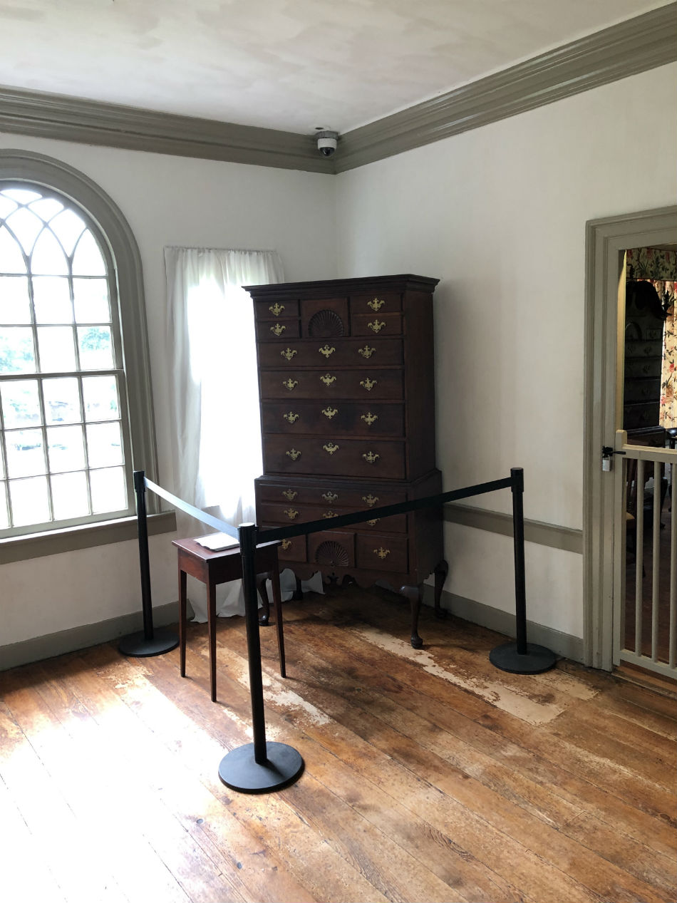 High Chest donated to Morristown NHP 75 years ago by Morristown Jewish Center. Picture of a dark wood high chest of drawers. The high chest is positioned in the corner of a room with white walls. There is a black stanchion around the chest.