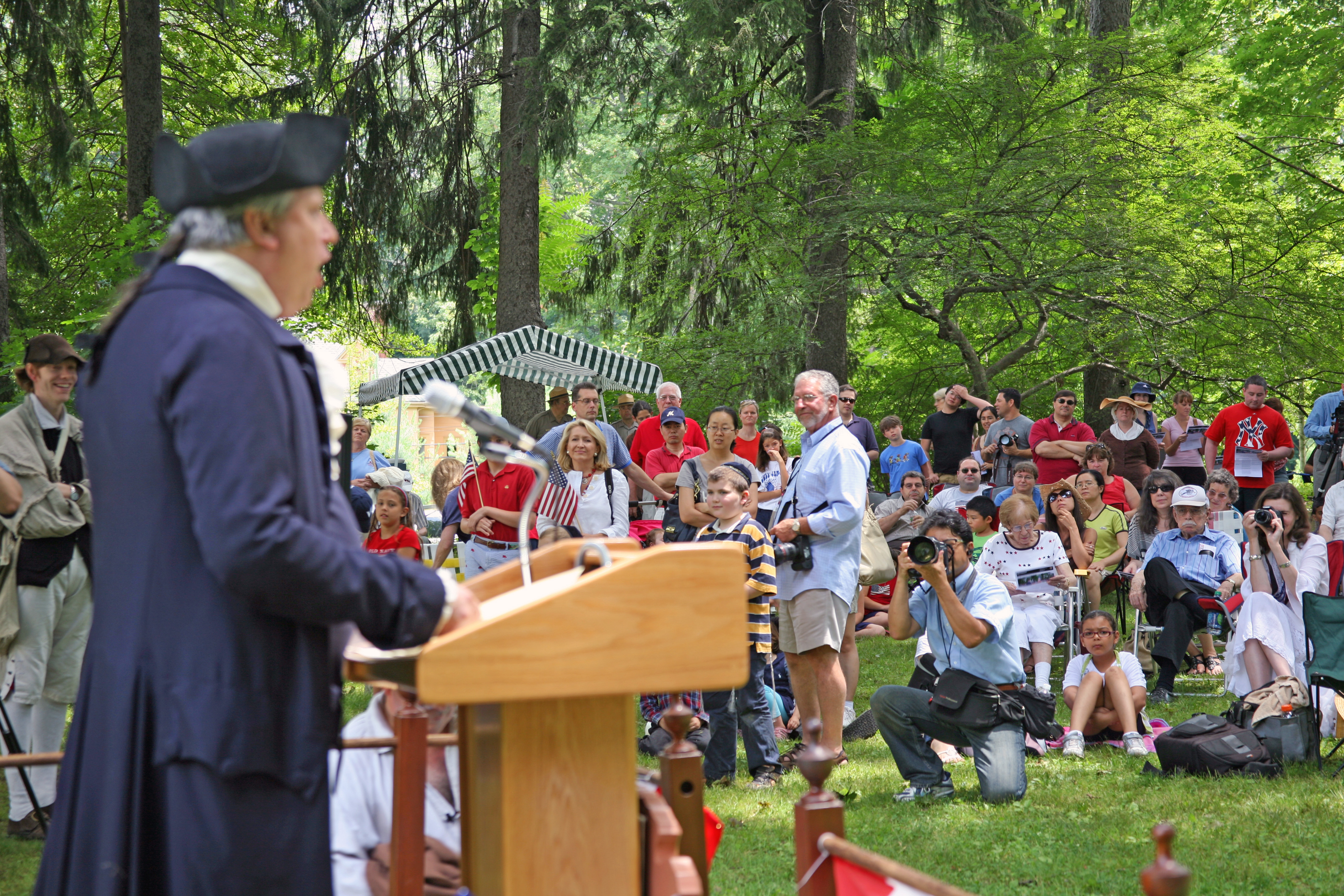 Reading of the Declaration - July 4, 2022 A speaker in colonial attire addresses a diverse crowd during an outdoor historical event. The audience includes children and adults, some holding American flags and taking photos, with a backdrop of trees and a canopy providing shade.