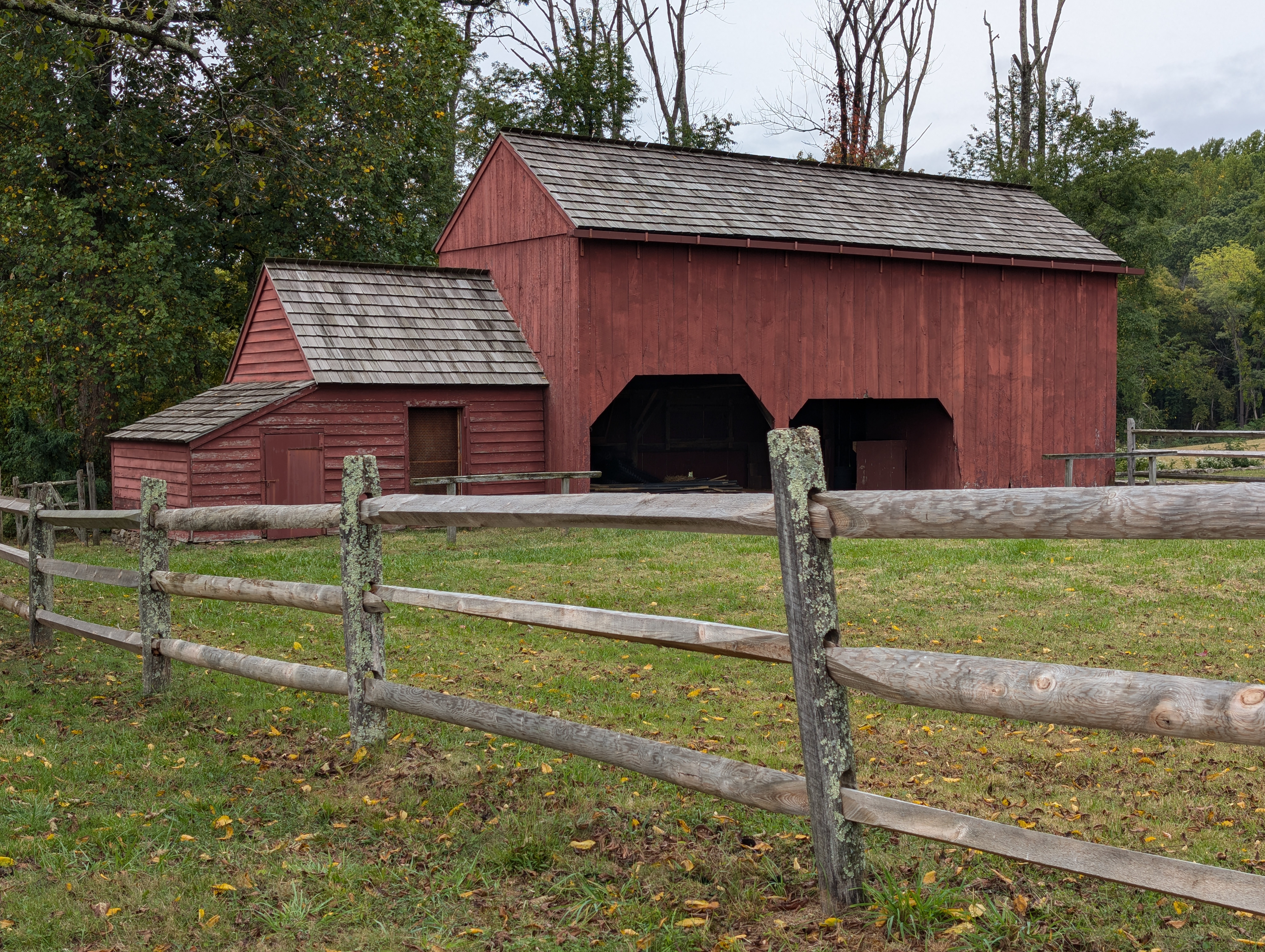 Wick Barn in September 2024 A red, wooden barn stands in the background, with green grass on the ground. Most of the trees surrounding the barn have green leaves on them, save for a couple that are devoid of leaves. A wooden fence stands in the foreground.