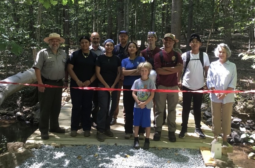 Park employees and partners getting ready to cut the ribbon on the newly restored bridge on the Grand Loop Trail. Twelve people standing on wooden bridge in the forest getting ready to cut the red ribbon to open the bridge to visitors.