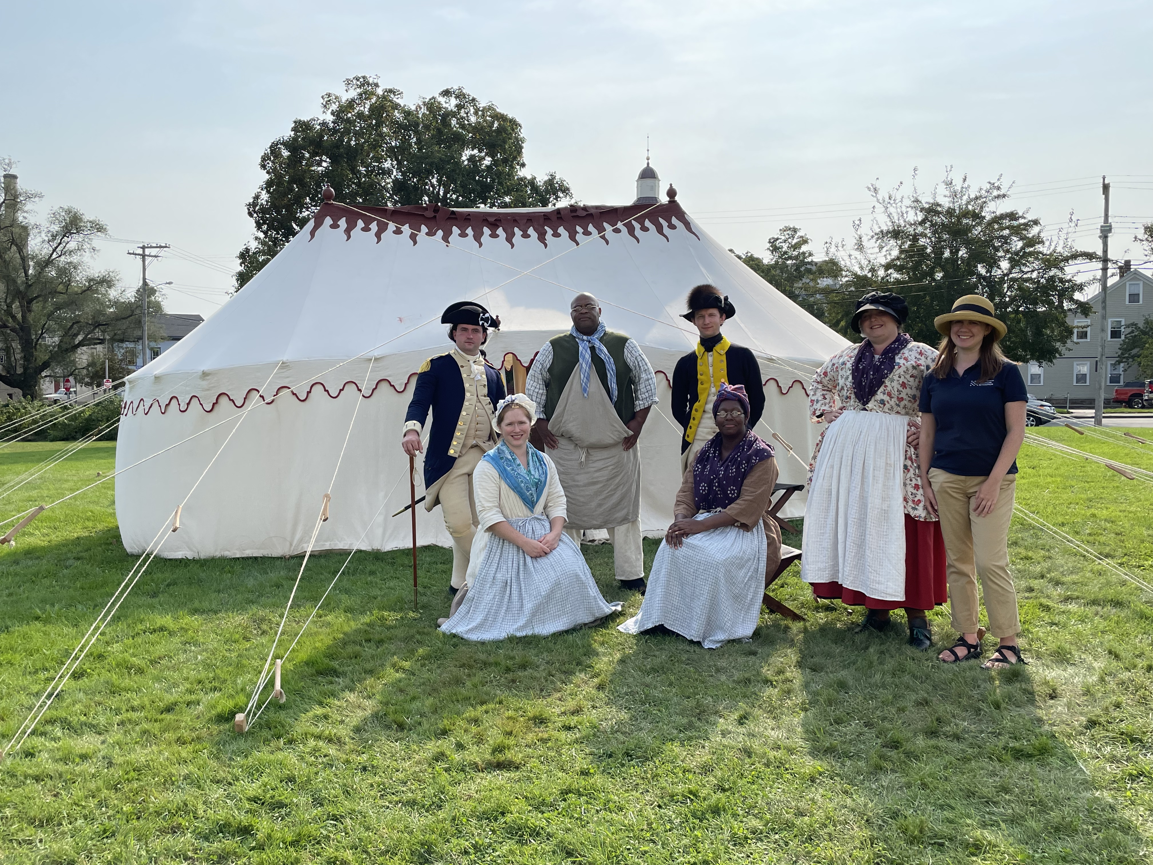 Seven reenactors in eighteenth century costumes standing in front of a replica of George Washington's  white oval tent with maroon trim on top.