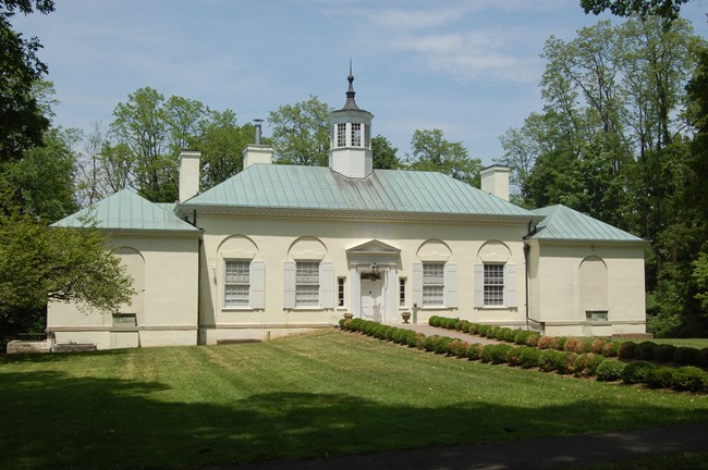 A large white museum building with green copper roof on a grassy lawn with a bush lined brick walkway