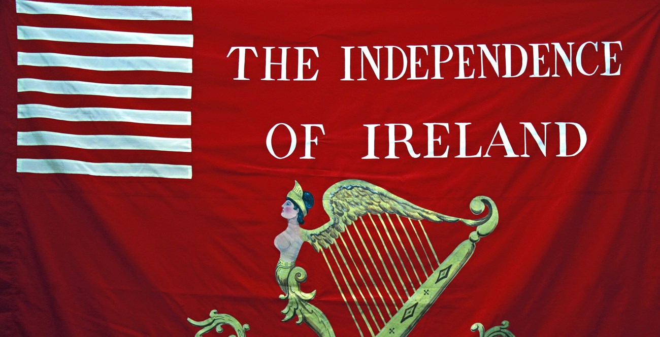 A large red flag, partially shown, with emblems of a stylized harp and white horizontal lines with the words "The Independence of Ireland"