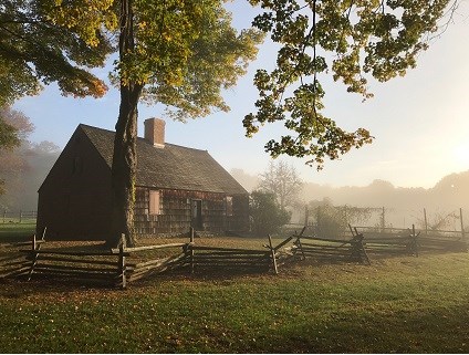 A two story Cape Cod farmhouse,, flanked by a split rail fence and a fenced in kitchen garden. A large oak tree towers above the home rising out of the frame. It is early morning in early fall, with sunlight breaking through the fog surrounding the house.