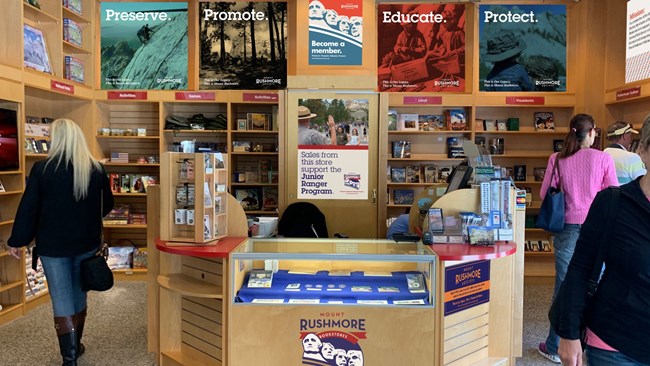 Information Center Bookstore with brown check out counter in center, brown shelves with bookstore merchandise, and four customers walking around.