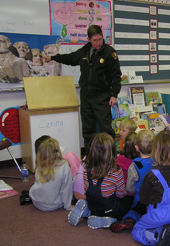 A Mount Rushmore park ranger wearing a green jacket and pants point to an image of Mount Rushmore.  Eight children sit on the floor with their backs to the photographer.