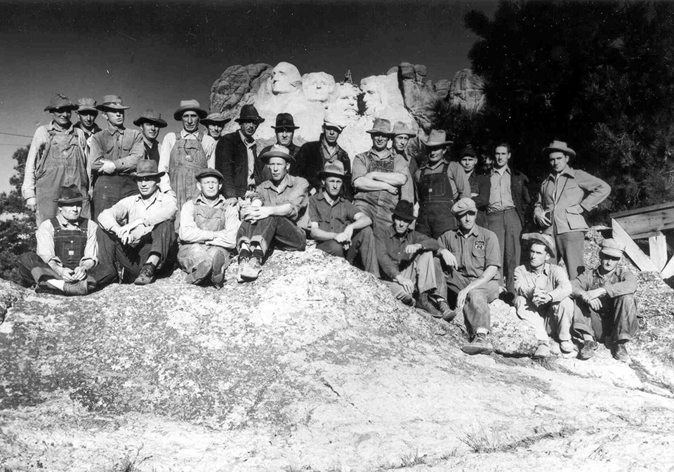 Black and white image of 24 workers in two rows, one seated, one standing on a large rock with Mount Rushmore in the background.  They are all white males wearing hats and clothing suitable for work - overalls, long sleeve shirts and boots.