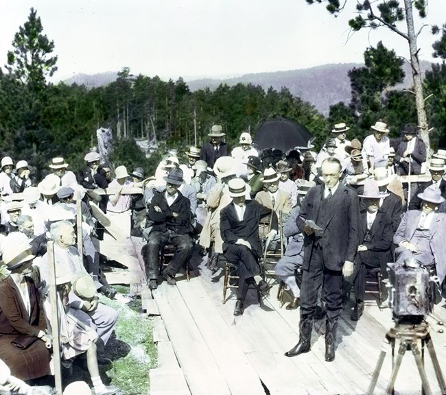 President Calvin Coolidge, wearing a dark suit and cowboy boots delivers a speech on August 10, 1927, at the Mount Rushmore dedication ceremony.  He is surrounded by a small crowd, some standing, some sitting, many wearing hats.