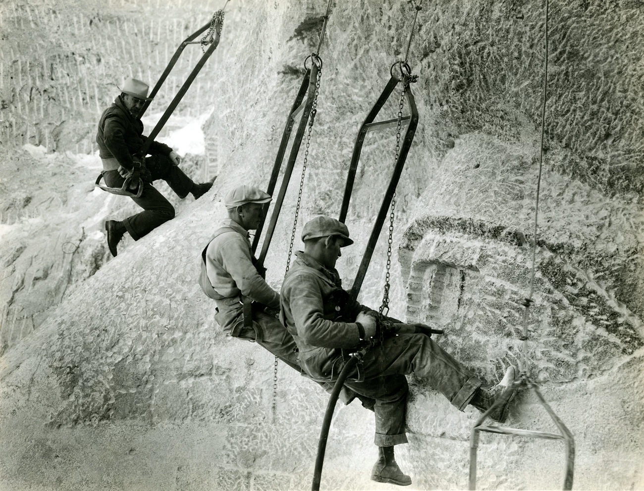 Gutzon Borglum observes two workers carving Jefferson's eye on Mount Rushmore. Gutzon Borglum on a sculpted nose in the upper left of the image observes two workers in the bottom center carving an eye on Mount Rushmore.  All 3 are in bosun's chairs.  The two workers are using pneumatic drills.