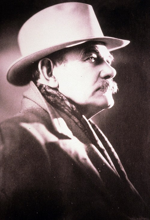 A black and white photograph of Gutzon Borglum, a white male with a mustache in his sixties, turned to the side and looking towards the right.  He wears a light fedora, a scarf and overcoat with the collar flipped up in the back.