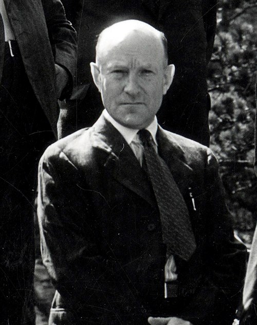 Black and white image of John Boland.  He is a balding white male in his forties looking straight at the photographer with a serious expression on his face.  He wears a dark suit, light-colored shirt and patterned necktie.