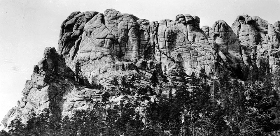 Foto in bianco e nero del Monte Rushmore prima dell'intaglio, con il suo aspetto rugoso e segnato dalle intemperie come l'avrebbe visto Charles Rushmore nel 1884-85.