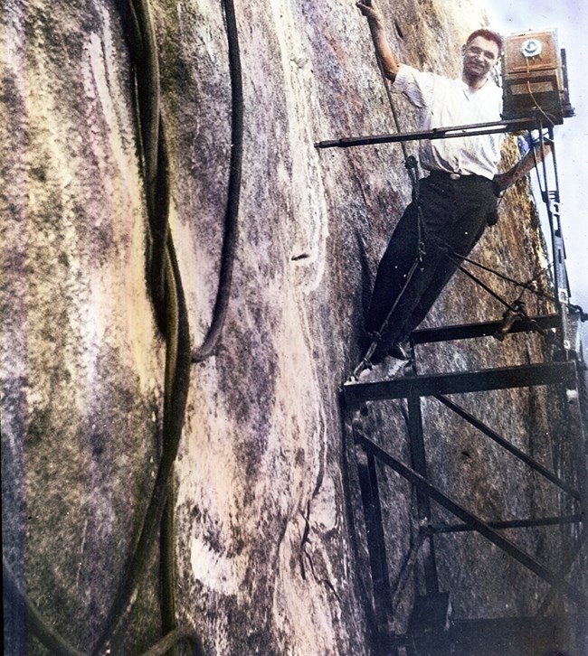 Photographer Charles D'Emery standing on metal scaffolding and posing with his camera on the side of Mount Rushmore.  Tangled air hoses run from the bottom to the top of the left side of the image.