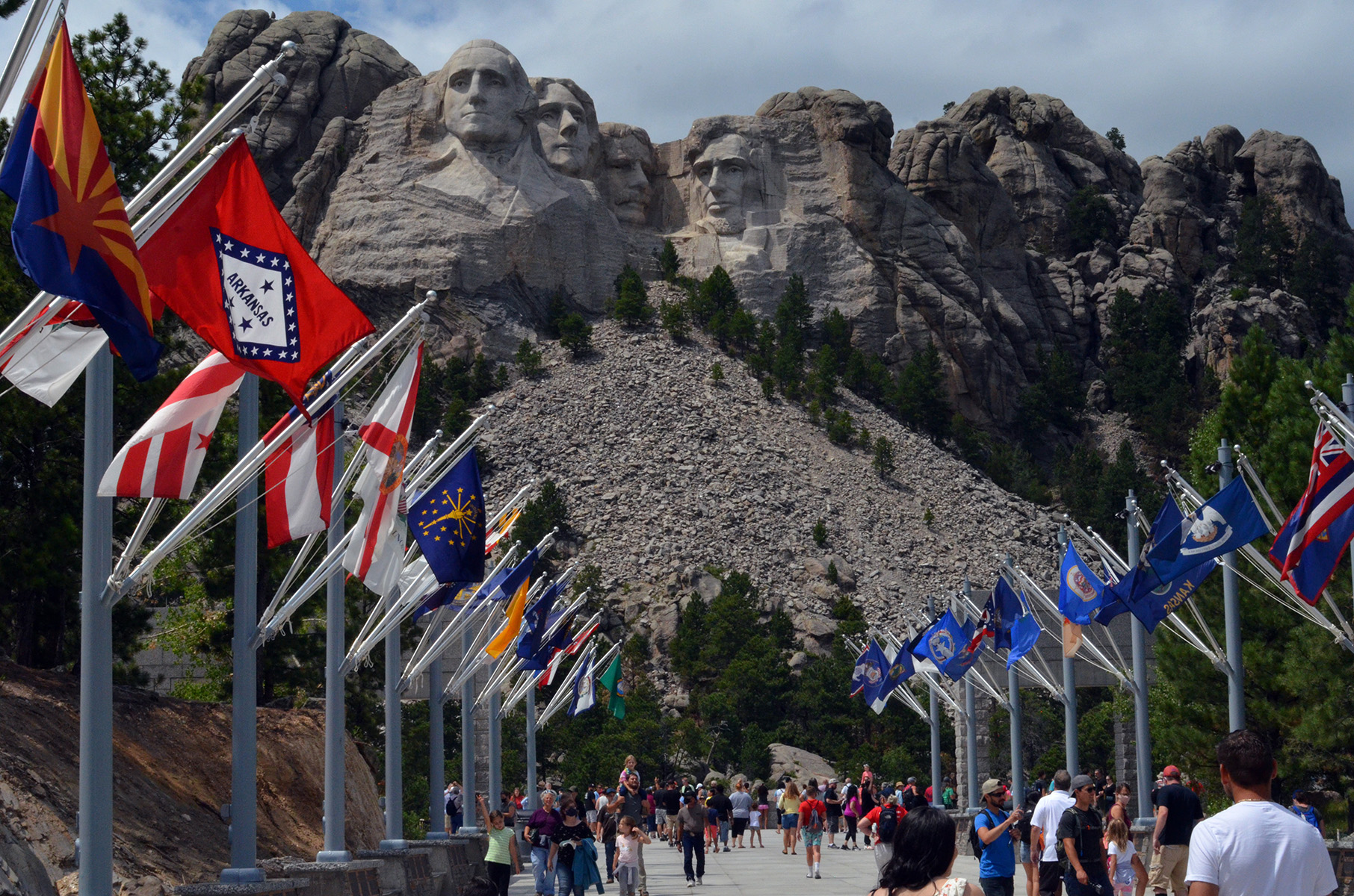 Avenue of Flags Four faces etched into side of grey mountain with numerous green trees. Hundreds of people walk on concrete walkway with two rows of USA state flags on either side.
