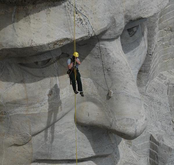 sculpture preservation rock block monitoring001 A person with a yellow hard-hat scaffolds off of a large grey rock sculpture of a face approximately 60’ tall.