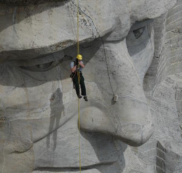 A person with a yellow hard-hat scaffolds off of a large grey rock sculpture of a face approximately 60’ tall.