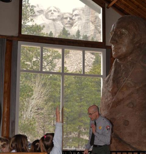 A uniformed park ranger delivers a program in front of numerous visitors of various ages. The Mount Rushmore sculpture is framed in a window behind him and he is standing next to a smaller replica of it.
