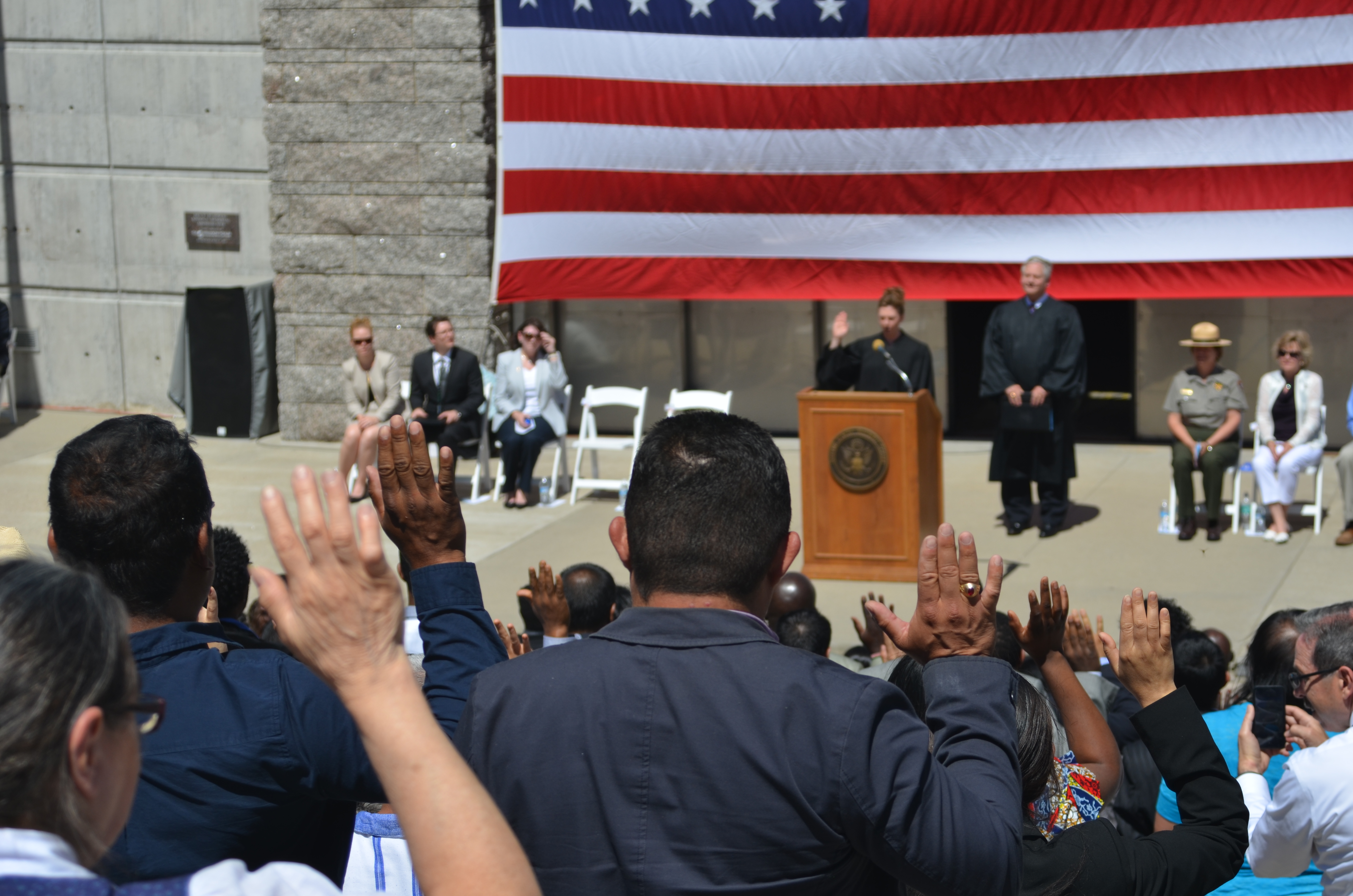 Naturalization Ceremony001 Crowd of people with right hand raised standing in front of stage with American flag prominently featured and a judge with right hand raised.