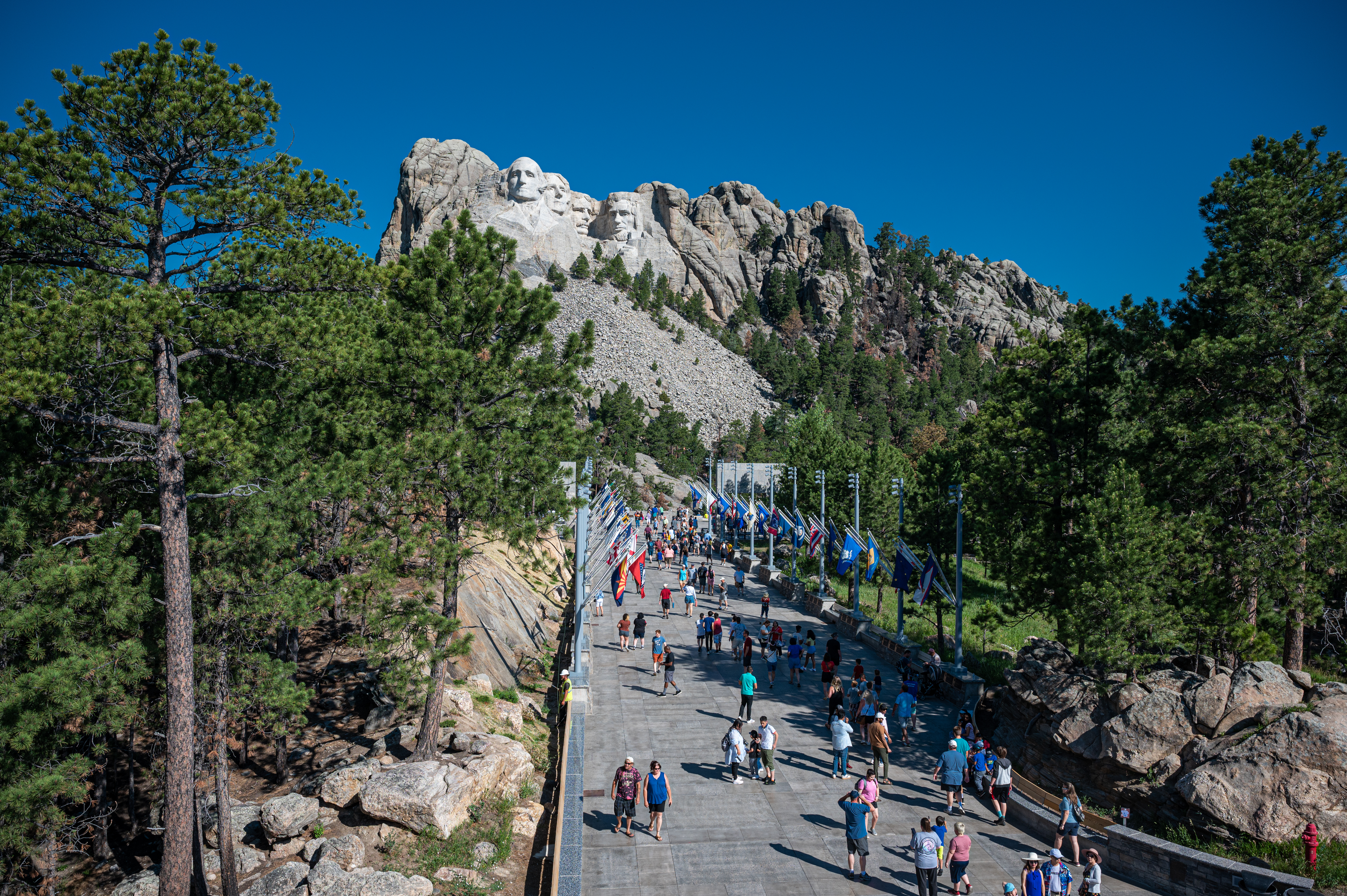 A concrete walkway with state flags on either side leading to Mount Rushmore. Dozens of people are on the walkway varying a variety of summer wear.