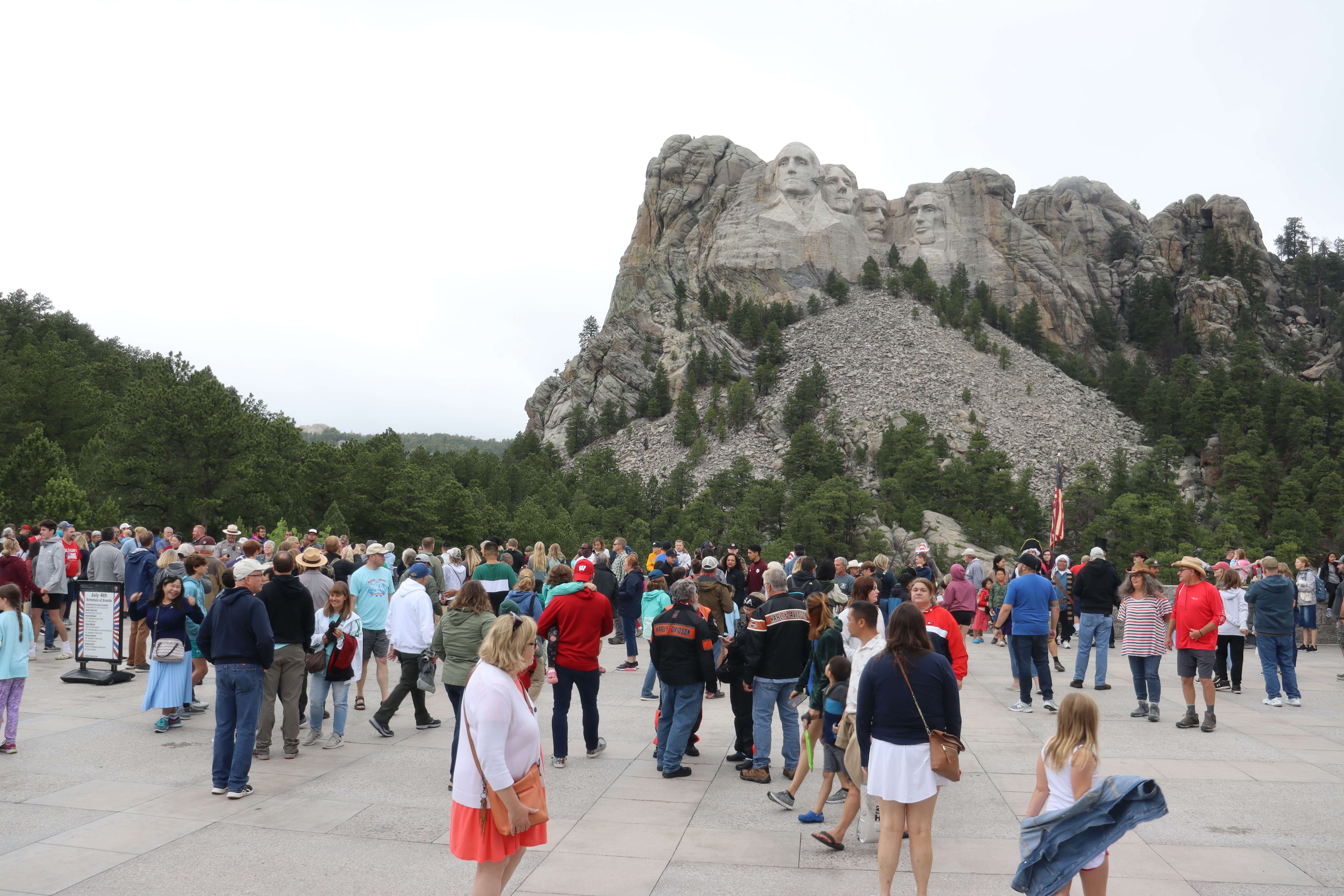 dozens of people standing in front of Mount Rushmore under a overcast sky.