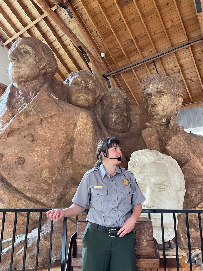 A uniformed park ranger with a microphone headset standing in front of a replica of Mount Rushmore.