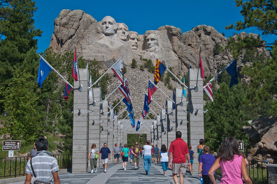 Visitors walk along the Avenue of Flags with Mount Rushmore in the background.