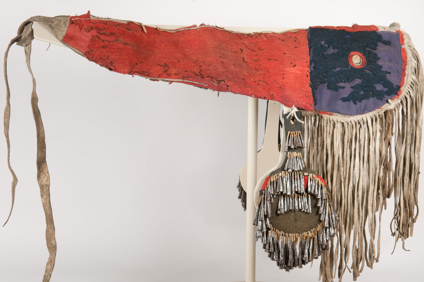 A cloth and bison hide horse crupper decorated with 'canteen' side pendents, jingles, and fringe. Each panel is made from scarlet and navy blue colored tradecloth. The tin cones, or 'jingles,' are made of pieces of tin, bent and rolled into shape. 