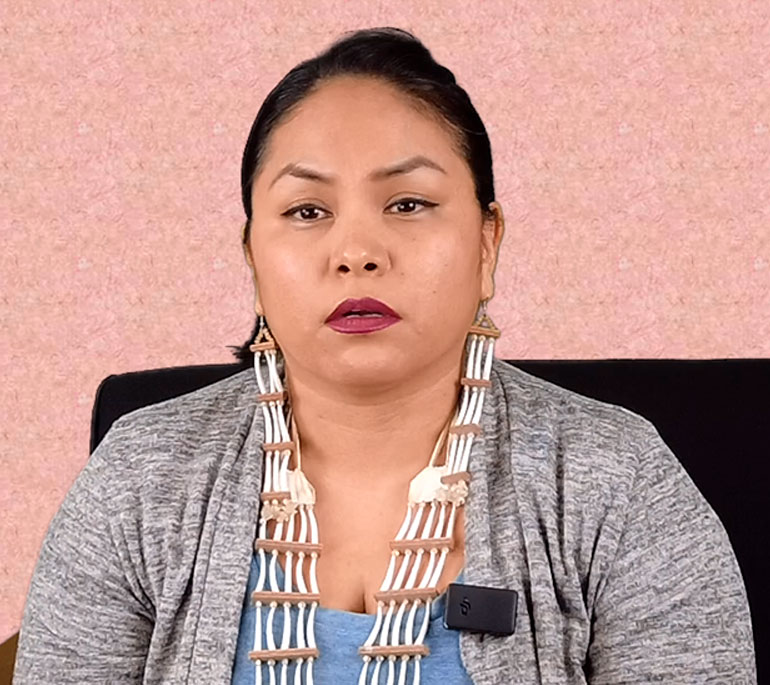 Native American woman sitting in a chair. She is wearing a gray shirt with a light blue tank top underneath and a necklace.