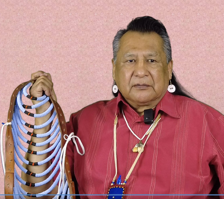 Native American man wearing a red collared shirt. He is holding a beaded piece.
