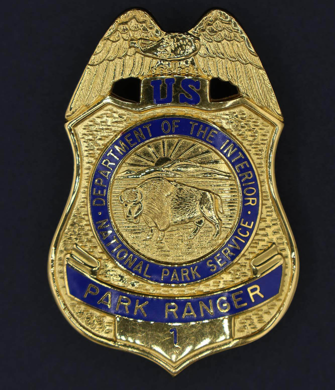 Gold shield-shaped badge with eagle on top. Marked U.S. Park Ranger and 1 in blue. Round seal has blue border with bison in the middle.