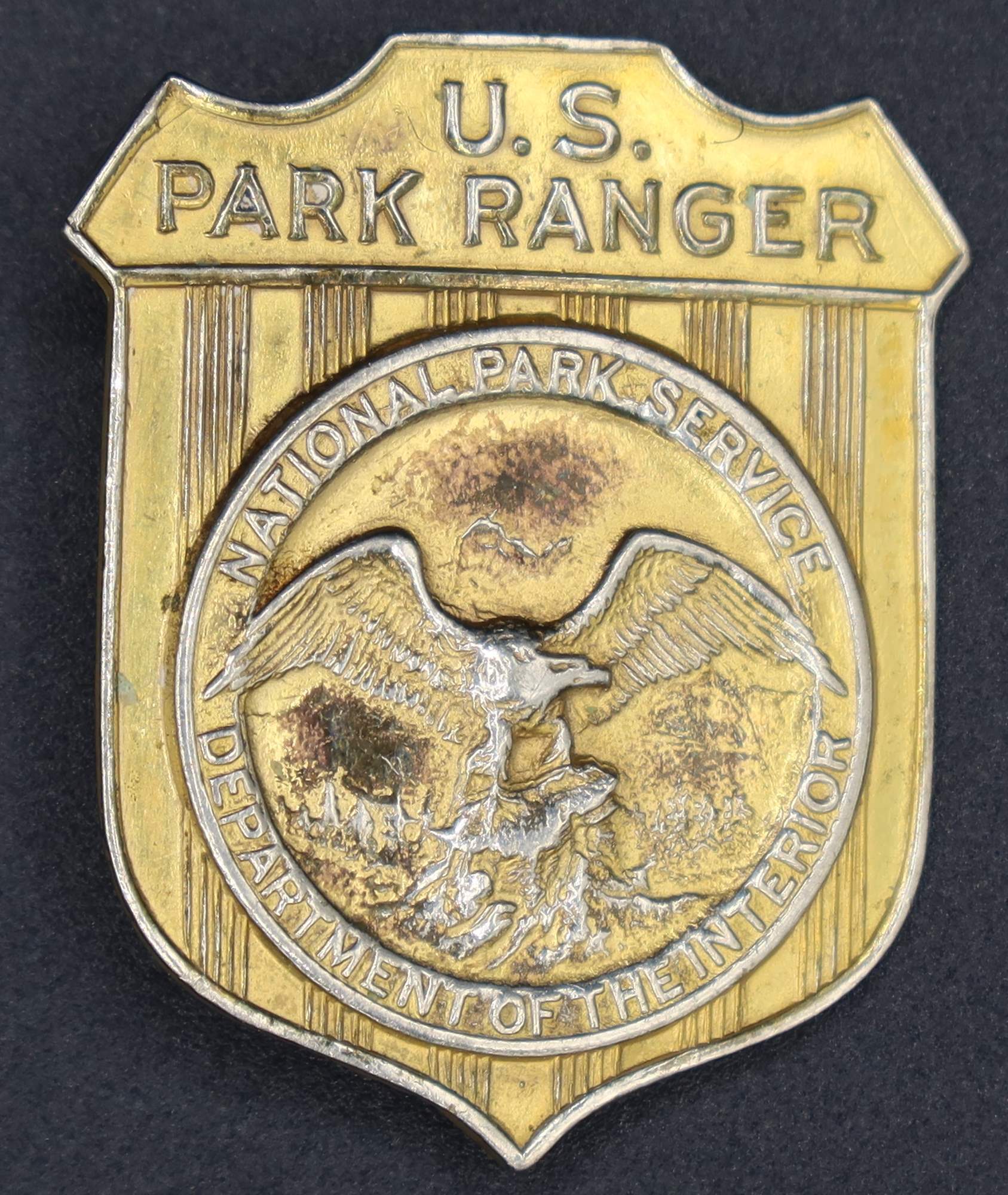 Gold shield-shaped badge marked U.S. Park Ranger. The raised round seal in the middle has an eagle looking to its left.