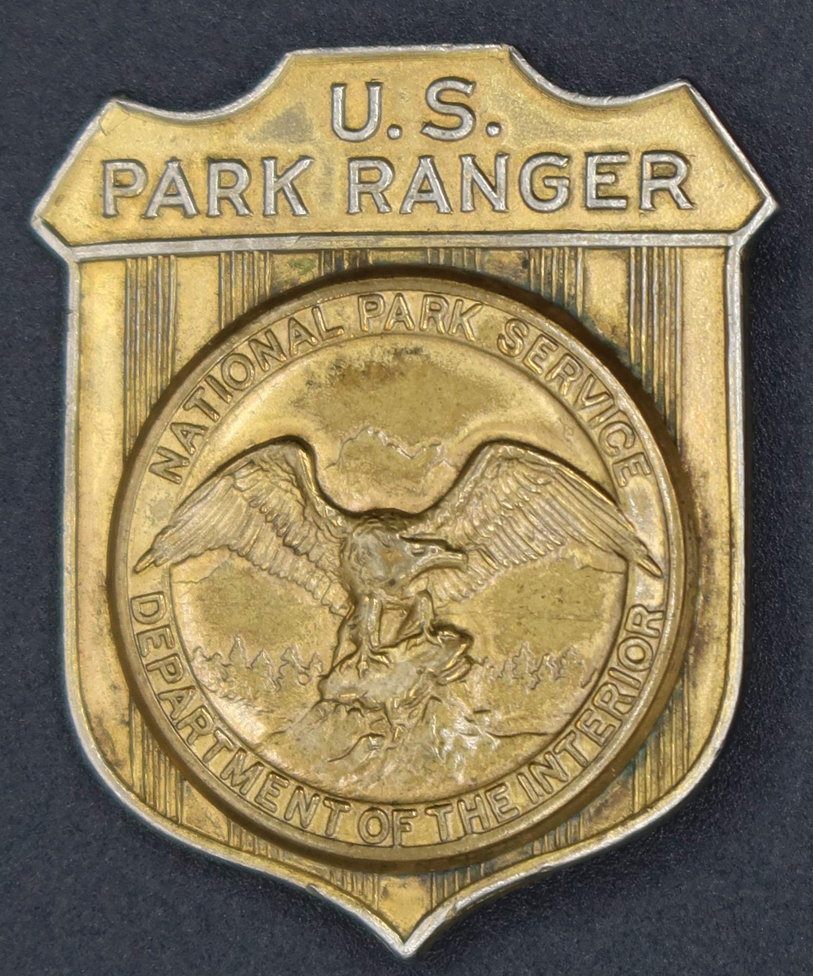 Gold shield-shaped badge marked U.S. Park Ranger. The raised round seal in the middle has an eagle looking to its left.