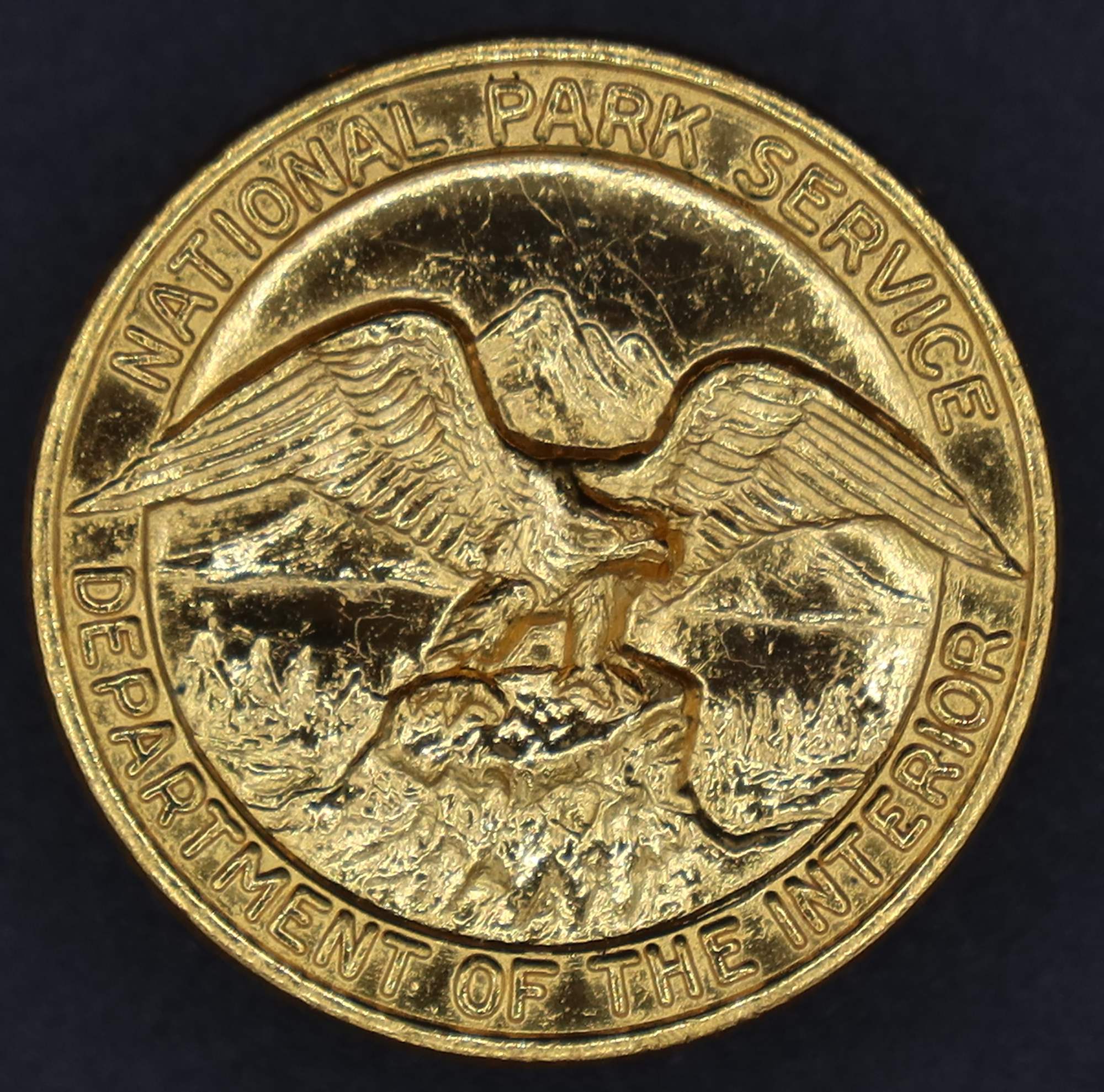 Round gold badge with National Park Service Department of the Interior around the rim and an eagle looking to its left in the middle.