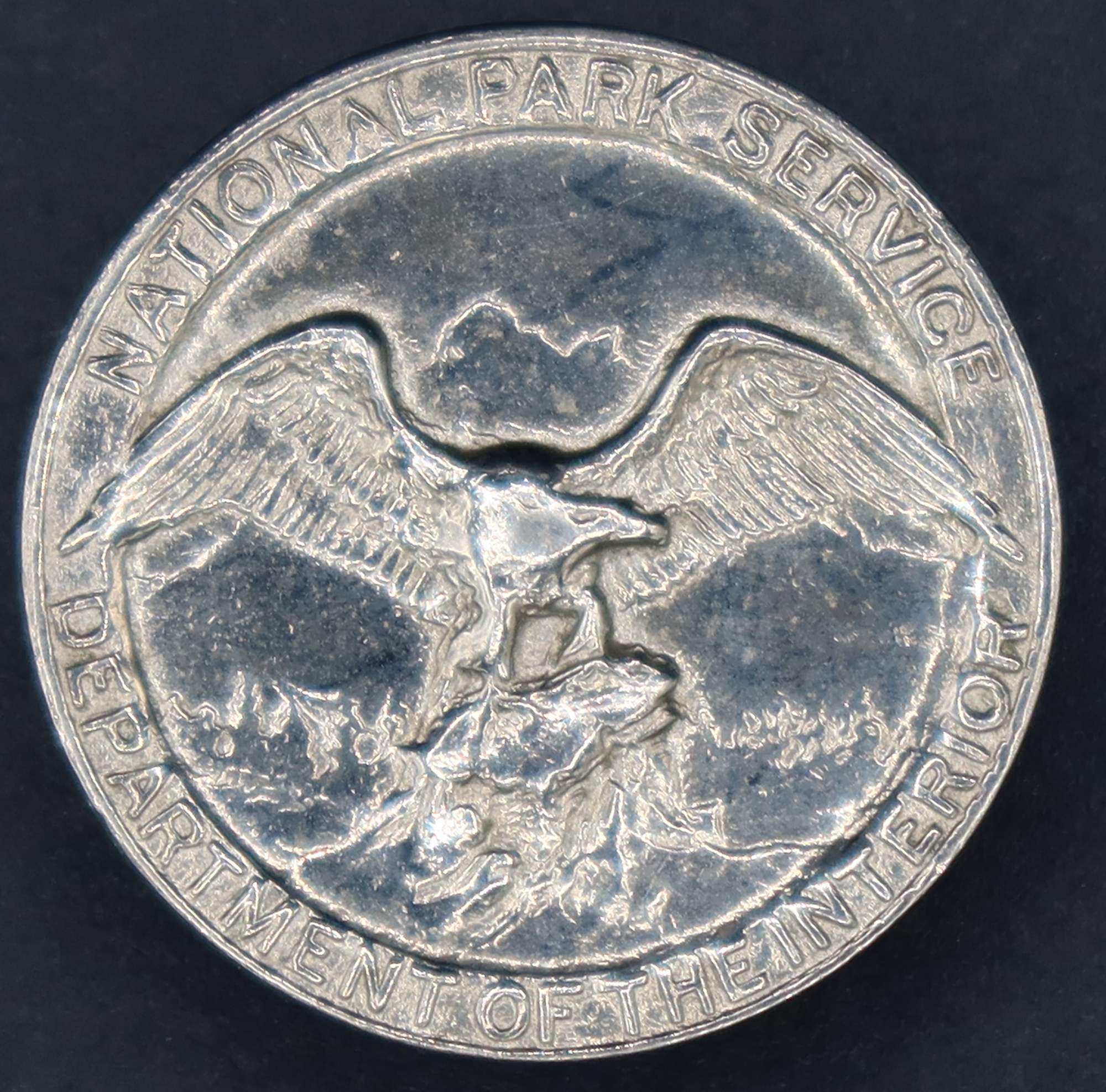 Round silver badge with National Park Service Department of the Interior around the rim and an eagle looking to its left in the middle.