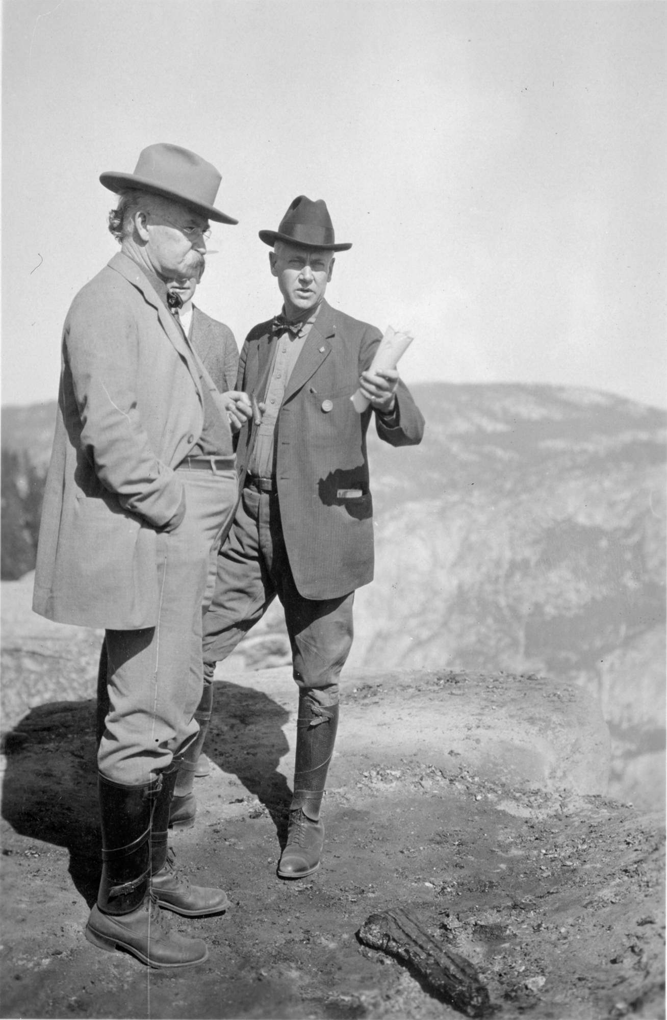 Director Stephen Mather, standing with two men, wears breeches, shoes, puttees, shirt, bow tie, dark cowboy hat, and a round badge on his coat.