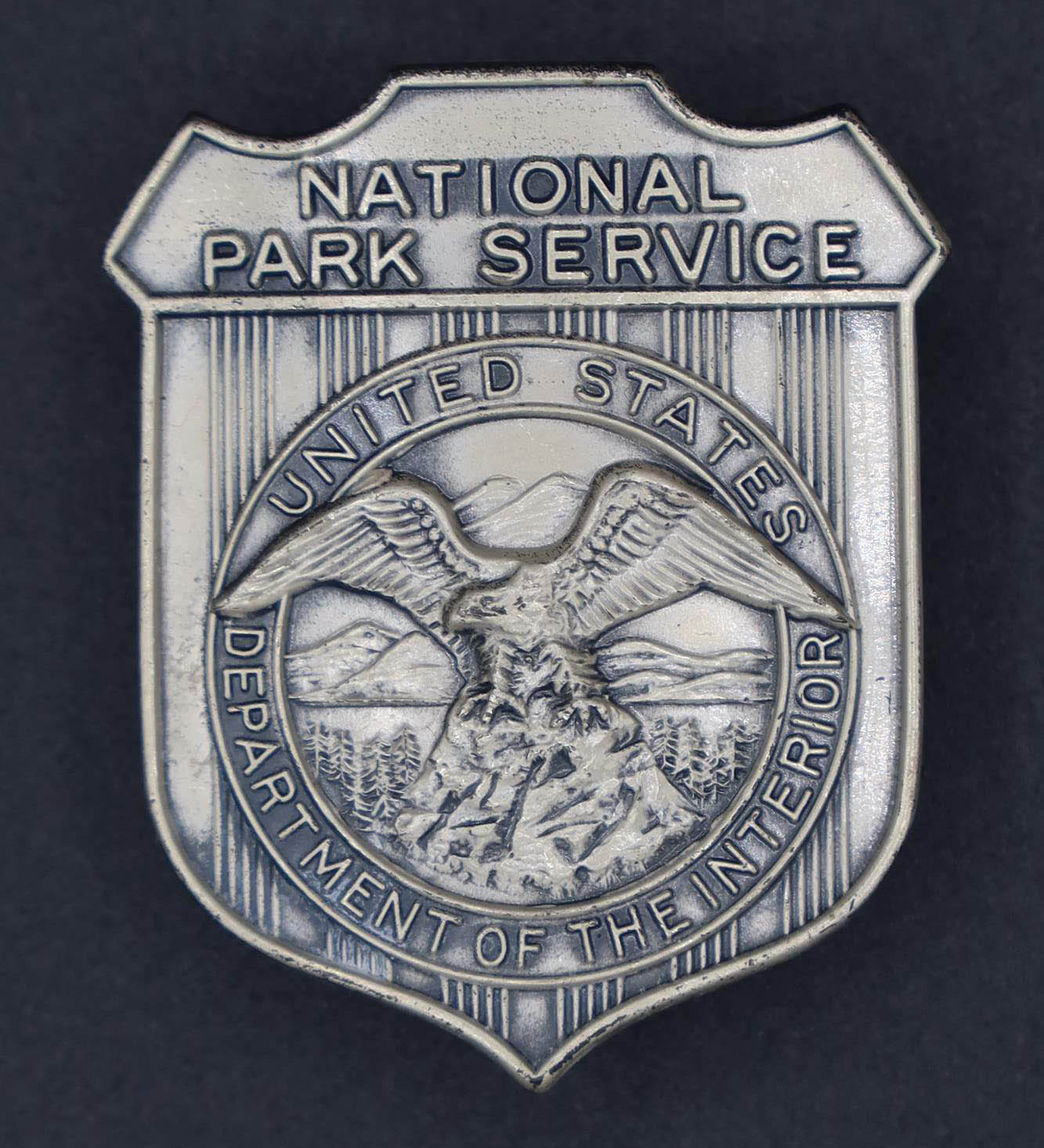 Silver shield-shaped badge marked National Park Service. The round seal in the middle has an eagle looking to its right.
