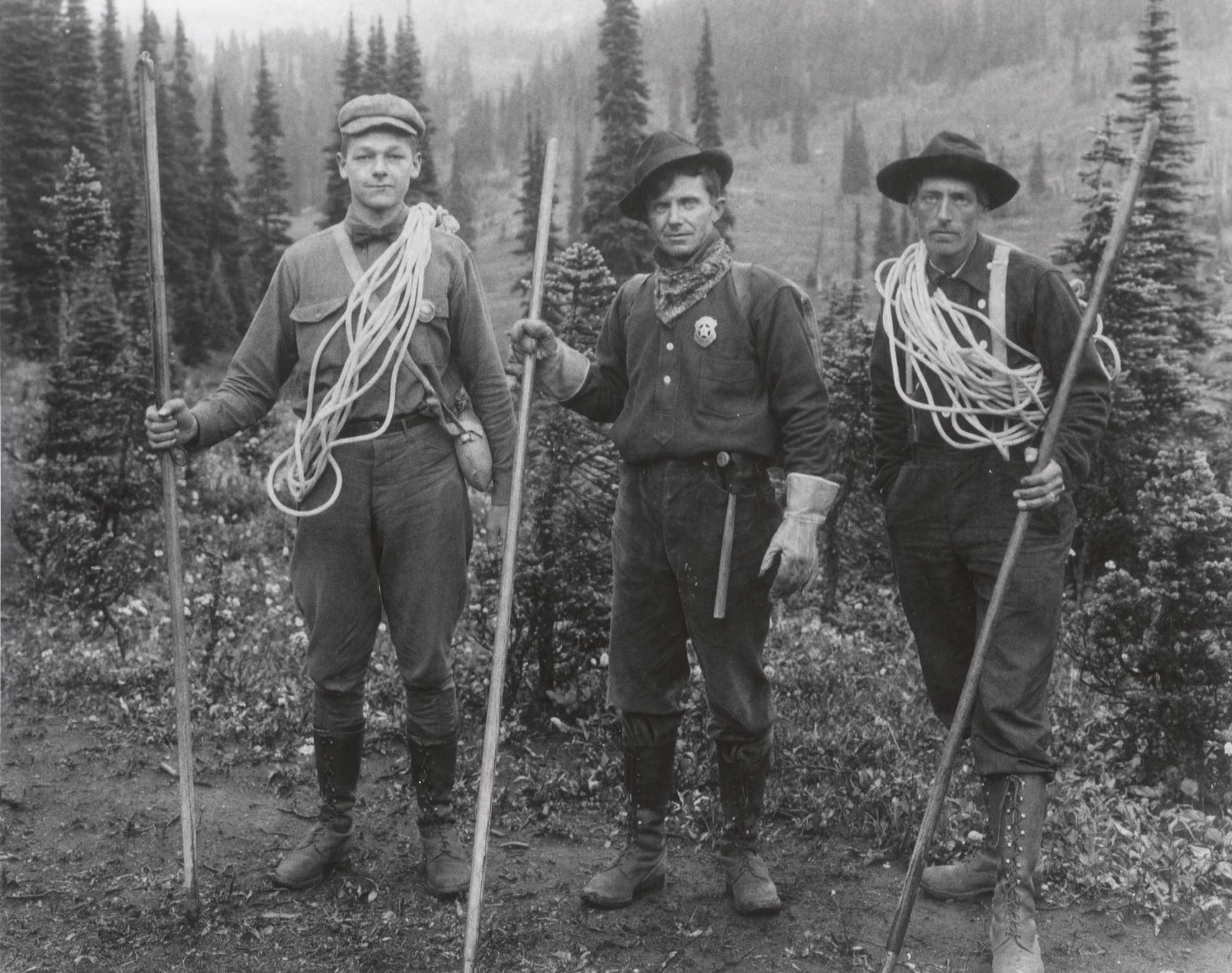 Phil Barrett, Joe Stampfler, and another man carry climbing ropes and walking sticks. Stampfler wears a shield-shaped badge and Barrett a round one.