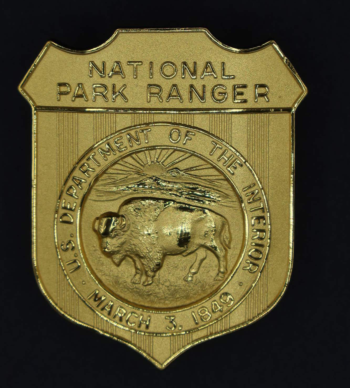 Gold shield-shaped badge marked National Park Ranger. Round seal in the middle has a bison with US Department of the Interior March 3, 1849 around it.
