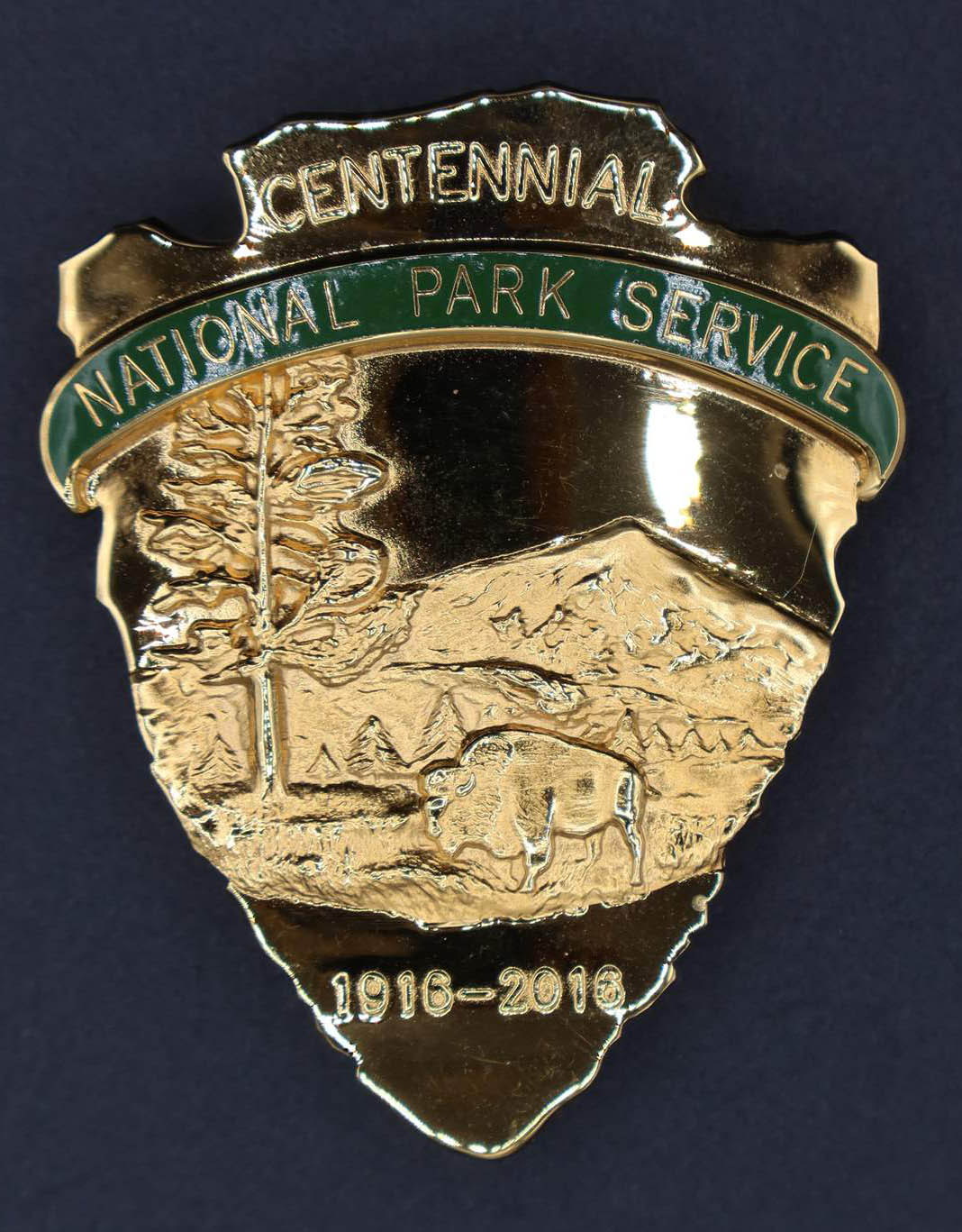 Gold arrowhead-shaped badge featuring bison scene engraved with Centennial at top and 1916-2016 below. Green banner reads National Park Service.