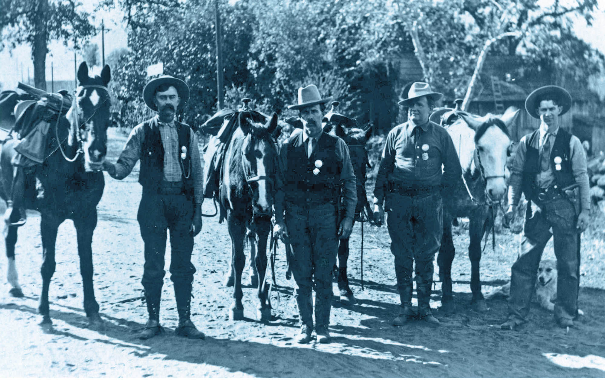 Four men in cowboy-type riding clothes stand next to horses. Each wears a round badge above a shield shaped badge, on a vest or shirt.  