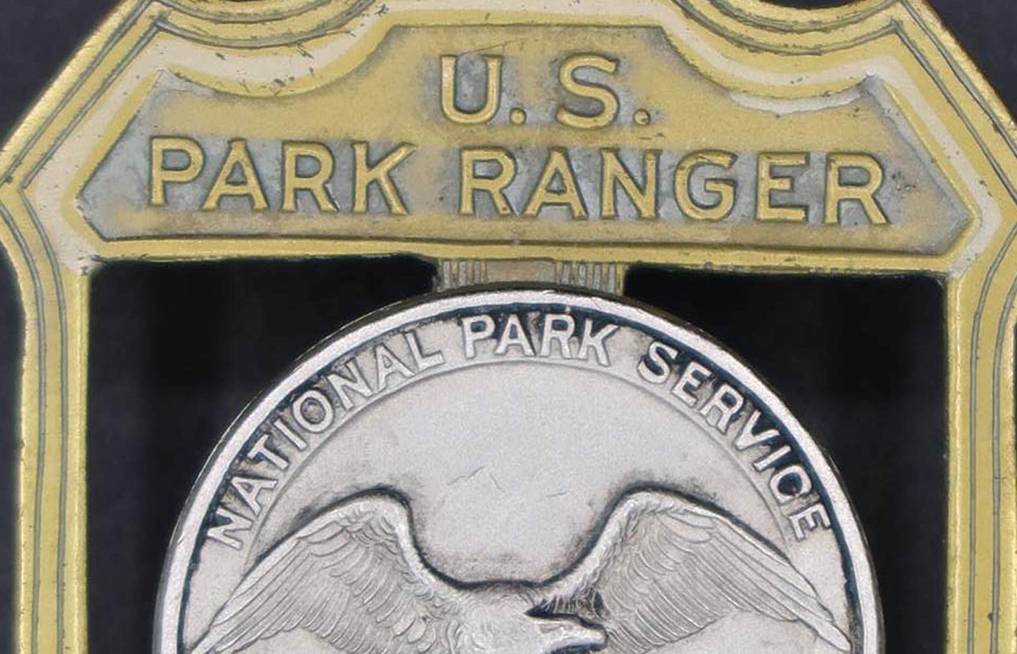 Brass shield-shaped U.S. Park Ranger badge. The raised round seal in the middle has an eagle looking to its left. Area around the center cut out and open.