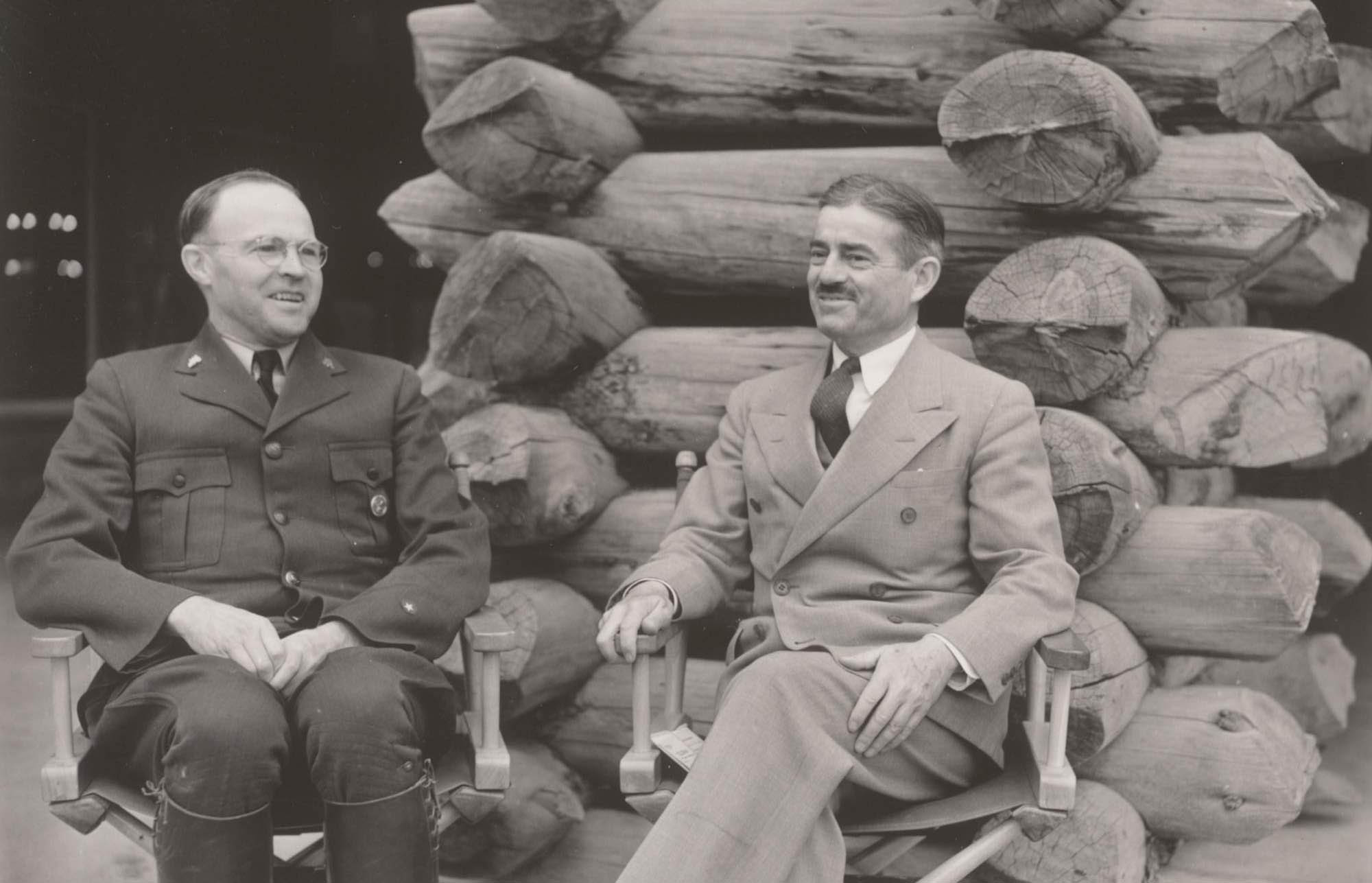 Edmund Rogers and Newton Drury sit in front of a stack of logs. Rogers wears the NPS uniform with a round badge. Drury wears a suit.
