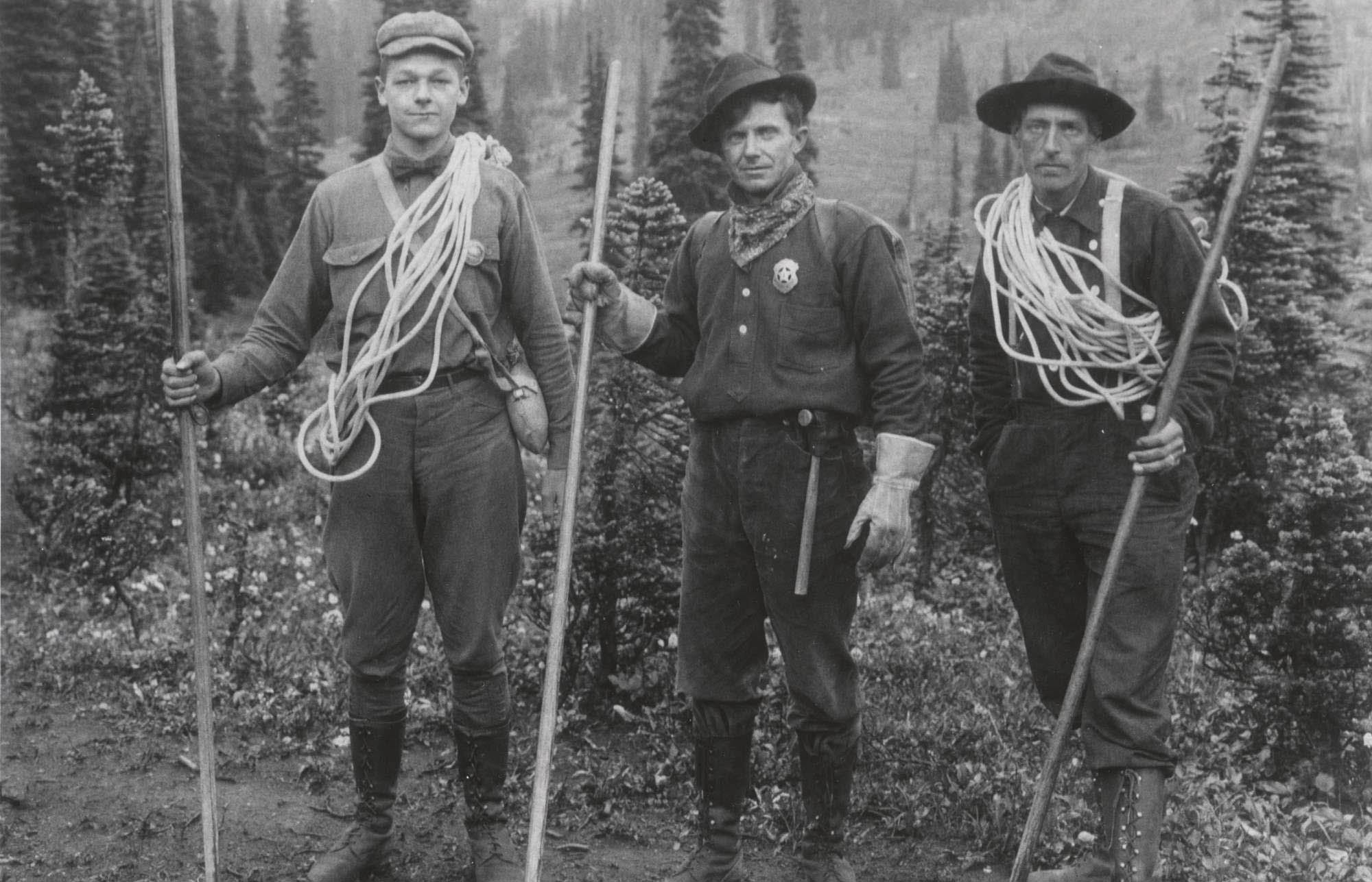 Phil Barrett, Joe Stampfler, and another man carry climbing ropes and walking sticks. Stampfler wears a shield-shaped badge and Barrett a round one.