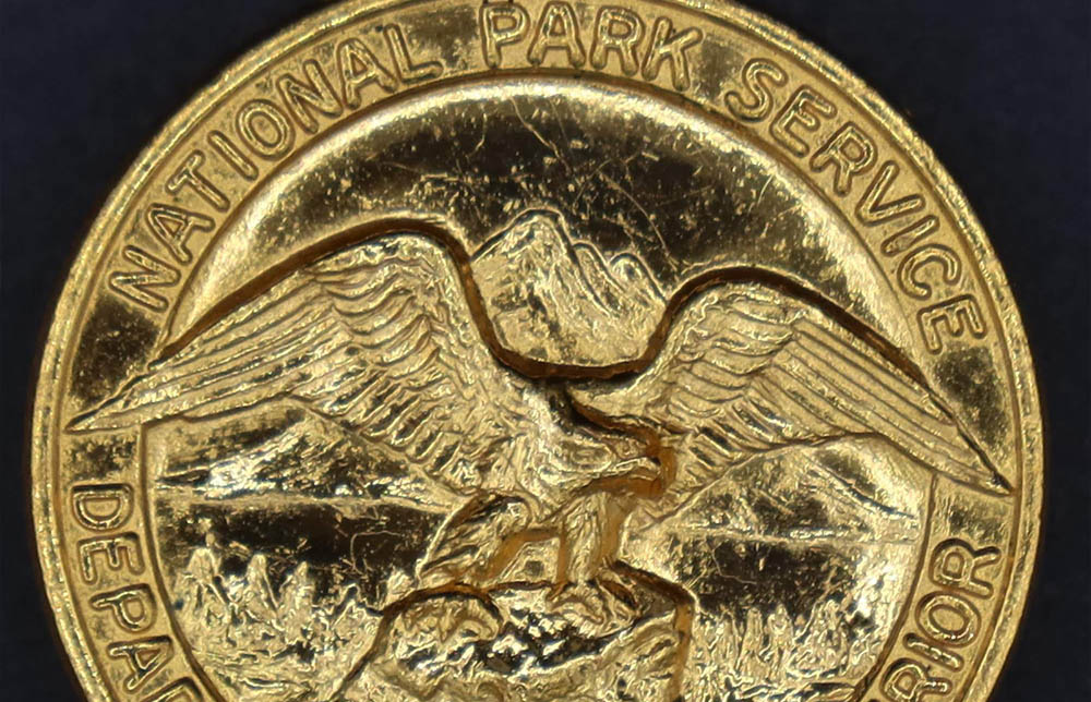 Round gold badge with National Park Service Department of the Interior around the rim and an eagle looking to its left in the middle.