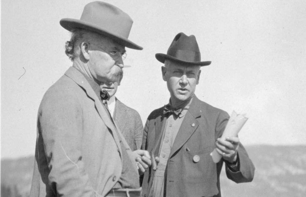 Director Stephen Mather, standing with two men, wears breeches, shoes, puttees, shirt, bow tie, dark cowboy hat, and a round badge on his coat.