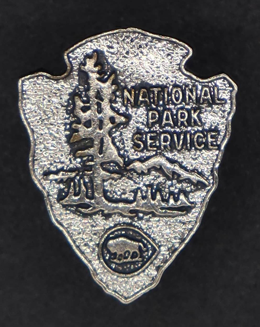 Silver arrowhead shaped pin with trees, mountains, bison, and words National Park Service.