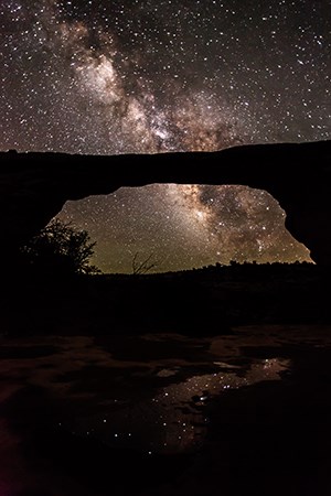 a massive stone arch with the milky way overhead. stars reflect in a pool of water at its base
